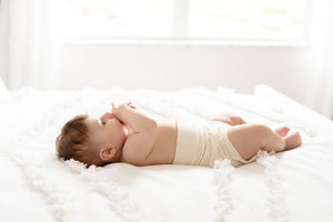 baby wearing naturelle unbleached cotton bloomer laying on the bed and eating fingers
